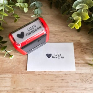 May include: A red and black self-inking stamp with the text "LUCY FINNEGAN" and a heart symbol. The stamp is used to create a black ink impression on a white card with the same text and heart symbol.