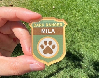 Könnte beinhalten: Ein schildförmiges Abzeichen in den Farben Grün und Braun. Der Text BARK RANGER ist oben in Gelb aufgedruckt, darunter steht der Name MILA in Weiß. Ein kreisförmiges Emblem mit einer braunen Pfotenabdruck-Grafik ist unten mittig platziert.