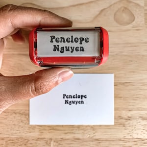 May include: A red and clear plastic self-inking stamp with the name "Penelope Nguyen" printed in black. The stamp is being used to stamp the name on a white card.