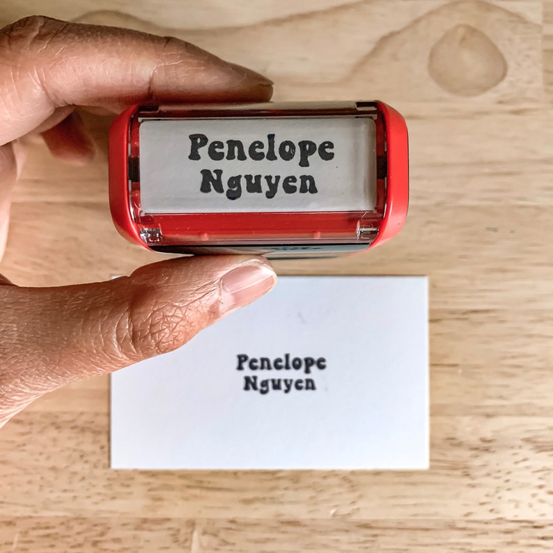 May include: A red and clear plastic self-inking stamp with the name "Penelope Nguyen" printed in black. The stamp is being used to stamp the name on a white card.
