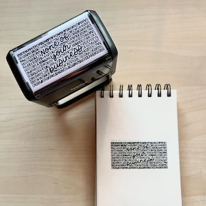 May include: A black rectangular stamp with the words "None of your business" in a stylized font. The stamp is next to a white notepad with the stamped phrase on it. The background is a light wood surface.