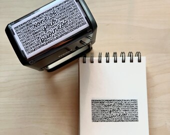 May include: A black rectangular stamp with the words "None of your business" in a stylized font. The stamp is next to a white notepad with the stamped phrase on it. The background is a light wood surface.
