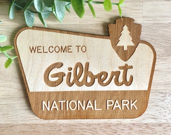 May include: Wooden sign with the text "Welcome to Gilbert National Park" in a retro style. The sign has a brown woodgrain finish and a white pine tree icon.