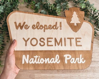 May include: A wooden sign with the text "We eloped! Yosemite National Park" in white lettering. The sign is shaped like a park sign with a stylized tree silhouette in the upper right corner.