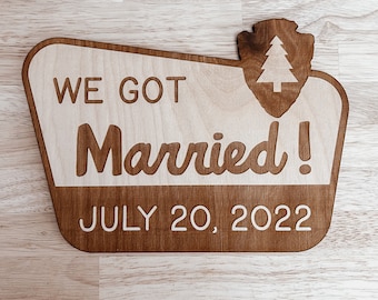 May include: Wooden sign with a rustic brown finish. The sign says "We Got Married! July 20, 2022". A small pine tree icon is in the upper right corner.