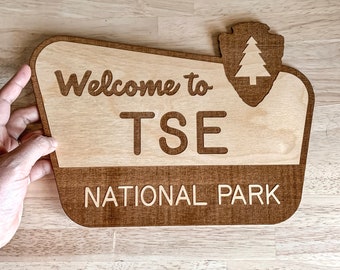 May include: Welcome to TSE National Park sign made of wood with a brown stain. The sign has a stylized tree and a park ranger symbol in the upper right corner.