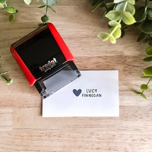 May include: A black and red self-inking stamp with the text "trodat printy 4711" on it. The stamp is used to create a black heart and the text "LUCY FINNEGAN" on a white card.