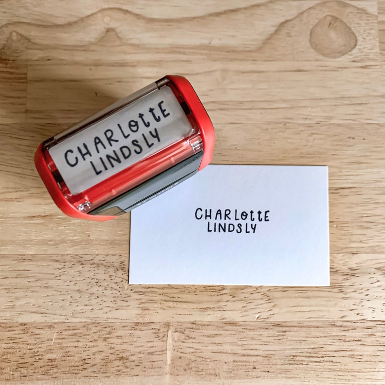May include: A red self-inking stamp with a white label that reads "CHARLOTTE LINDSLY". The stamp is next to a white card with the same text printed on it.