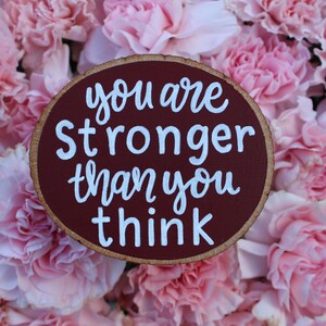 K&ouml;nnte beinhalten: Ein rundes Holzschild mit einem mahagonifarbenen Hintergrund und wei&szlig;en Buchstaben. Das Schild tr&auml;gt die Aufschrift "You are stronger than you think." Das Schild ist von rosa Nelken umgeben.