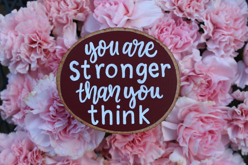 K&ouml;nnte beinhalten: Ein rundes Holzschild mit einem mahagonifarbenen Hintergrund und wei&szlig;en Buchstaben. Das Schild tr&auml;gt die Aufschrift "You are stronger than you think." Das Schild ist von rosa Nelken umgeben.