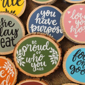 K&ouml;nnte beinhalten: Eine Sammlung runder Holzscheiben mit handgemalten inspirierenden Botschaften in verschiedenen Farben, darunter Gr&uuml;n, Blau, Rosa, Gelb, Orange und Lila. Die Botschaften lauten: "L&auml;cheln", "Sei stolz auf das, was du bist", "Du hast einen Sinn", "Du bist st&auml;rker, als du denkst", "Ich liebe dich mehr", "Wachse weiter", "Konzentriere dich auf das Gute" und "Nimm dir einen Tag zum Essen".
