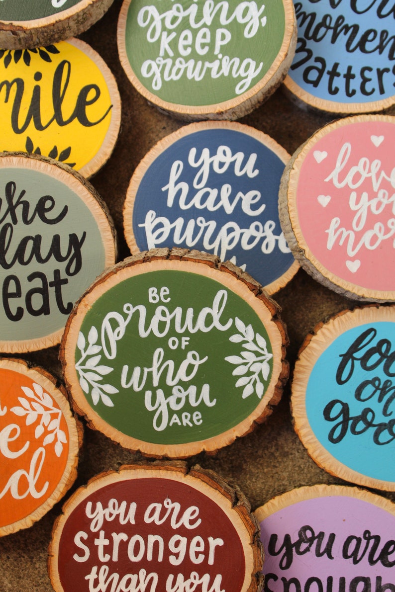 K&ouml;nnte beinhalten: Eine Sammlung runder Holzscheiben mit handgemalten inspirierenden Botschaften in verschiedenen Farben, darunter Gr&uuml;n, Blau, Rosa, Gelb, Orange und Lila. Die Botschaften lauten: "L&auml;cheln", "Sei stolz auf das, was du bist", "Du hast einen Sinn", "Du bist st&auml;rker, als du denkst", "Ich liebe dich mehr", "Wachse weiter", "Konzentriere dich auf das Gute" und "Nimm dir einen Tag zum Essen".