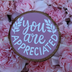K&ouml;nnte beinhalten: Ein rosafarbenes Holzschild mit den Worten "you are appreciated" in wei&szlig;er Farbe. Das Schild ist von rosa Nelken umgeben.