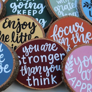 K&ouml;nnte beinhalten: Zehn runde Holzscheiben mit bemalten inspirierenden Botschaften. Die Botschaften beinhalten "You have purpose", "You are stronger than you think", "Enjoy the little things", "Focus on the good", "Keep going", "Just be you", "With grateful heart" und mehr.
