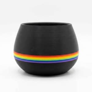 May include: A matte black, rounded ceramic planter or bowl featuring a horizontal rainbow-colored stripe encircling the lower section. The vessel has a wide, open top and a smooth, curved exterior surface with a consistent dark finish throughout.