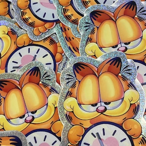 May include: A set of colorful stickers featuring Garfield the cat holding a clock with a heart on the face. The stickers have a glittery, iridescent finish.