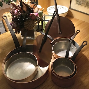 May include: A set of copper cookware with black handles. The set includes a large pot, a smaller pot, a saucepan, and two lids. The cookware is arranged on a wooden surface.