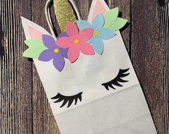 May include: White paper gift bag decorated as a unicorn. The bag features a gold glitter horn, pink, purple, and blue paper flowers, and black eyelashes. The bag has white handles and is set against a wooden background.