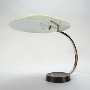 May include: A table lamp featuring a wide, circular, off-white shade with a white underside. The lamp has a curved, polished chrome arm that connects to a round, dark brown base. A small white power switch is located on the top of the base.