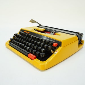 May include: A yellow vintage typewriter with black keys and a red button. The typewriter says "PRESTIGE DELUXE 230TR" on the front.