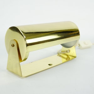 May include: A gold-toned metal desk lamp with a cylindrical shade and a white cord with a switch. The lamp has a simple, minimalist design.