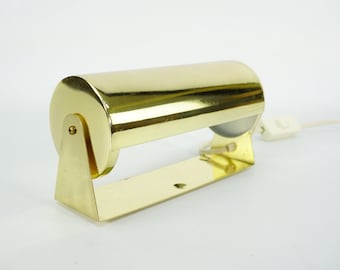 May include: A gold-toned metal desk lamp with a cylindrical shade and a white cord with a switch. The lamp has a simple, minimalist design.