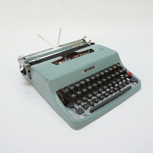 May include: A vintage Lettera 32 typewriter in a muted light blue finish. The device features a QWERTY keyboard with black circular keys, a prominent red key on the right side, a metal carriage assembly, and a silver nameplate on the front casing.