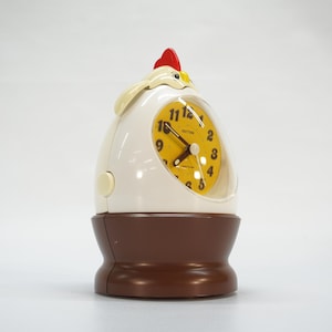 May include: A novelty alarm clock shaped like a chicken. The clock has a white egg-shaped body, a brown base, and a red comb. The clock face is yellow with black numbers and hands. The clock is approximately 6 inches tall.