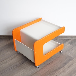 May include: A rolling side table featuring a bright orange frame with two white shelves. The structure includes a lower storage compartment and an open upper surface, supported by four small metal casters attached to the base for mobility.