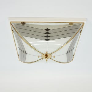 Könnte beinhalten: Eine geometrische Deckenleuchte mit weißem Rahmen und goldfarbenen Zierleisten. Die Glasscheiben weisen ein Muster aus grauen und schwarzen Formen auf. Die Leuchte ist an einer weißen Decke montiert.