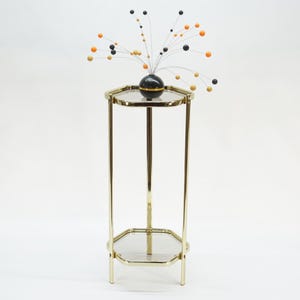 May include: A gold-toned metal plant stand with a hexagonal top and base. The stand supports a black spherical vase with a decorative arrangement of wire stems and colorful spherical accents in orange, black, and gold.