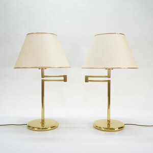 May include: A pair of brass table lamps with cream-colored lampshades. The lamps have a minimalist design with a circular base and a straight stem. The lampshades are slightly tapered and have a gold trim.