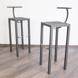 Op de afbeelding: Twee grijze metalen barkrukken met vierkante zittingen en gebogen rugleuningen. Elke kruk heeft een voetensteun en een minimalistisch ontwerp. De krukken staan op een houten vloer tegen een witte muur.