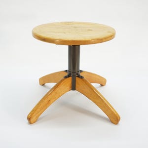 Op de afbeelding: Een vintage houten kruk met een ronde zitting en drie uitlopende poten. De zitting is licht van kleur en de poten zijn van hout. De centrale steun is donker metaal. De kruk is ontworpen om op te zitten.