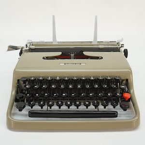 May include: A vintage typewriter with a light tan body, black keys, and a red button. The keys are arranged in a QWERTY layout. Two white paper guides stand upright. The typewriter is on a white background.