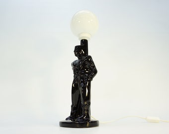 May include: Black ceramic table lamp with a white globe shade. The lamp base is shaped like a standing figure.