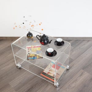以下が含まれることがあります： 透明なアクリル製のティーカート。2段の棚があり、黒いティーポット、2つのティーカップ、雑誌が置かれています。装飾的な茎が付いた黒い花瓶が上にあります。カートには移動しやすい小さな車輪が付いています。