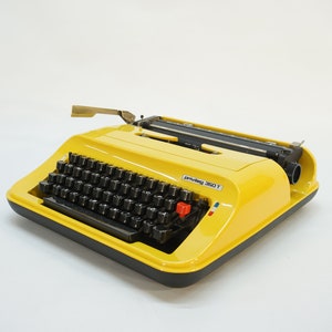 May include: A yellow vintage Olivetti Lettera 350 T typewriter with a black keyboard and a red button. The typewriter has a black and white French flag sticker on the side.