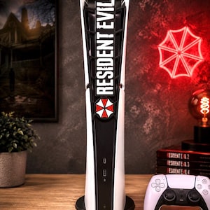 Pode incluir: Uma consola de jogos branca e preta com um logótipo vertical Resident Evil e um emblema vermelho e branco da Umbrella Corporation no painel frontal, acompanhada por um comando sem fios a condizer em branco e preto com botões e joysticks.