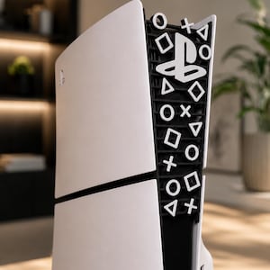 Könnte beinhalten: Eine weiße Spielkonsole mit einem schwarzen Mittelteil, der mit erhabenen weißen PlayStation-Symbolen wie Kreis, Kreuz, Quadrat und Dreieck sowie dem PlayStation-Logo am oberen Rand des schwarzen Bereichs verziert ist.