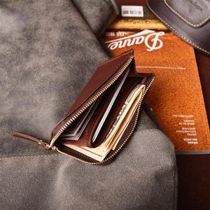 以下が含まれることがあります： 茶色の革製財布。ファスナー開閉式で、一部が開いており、現金とカードが見える。財布は灰色の表面に置かれ、背景には雑誌と革製品がある。雑誌には「Danne」の文字が書かれている。