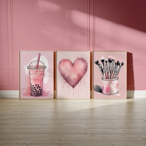 Op de afbeelding: Drie ingelijste aquarelprints met roze thema's. Eén toont een bubble tea, een andere een hart en de derde een verzameling make-up kwasten in een decoratieve container. De prints staan tegen een roze muur.