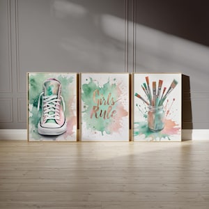 Op de afbeelding: Drie ingelijste aquarelprints. De eerste toont een high-top sneaker, de tweede luidt "Girls Rule" en de derde toont penselen in een pot. Elke print heeft een gouden lijst en een kleurrijke, gespatte achtergrond.