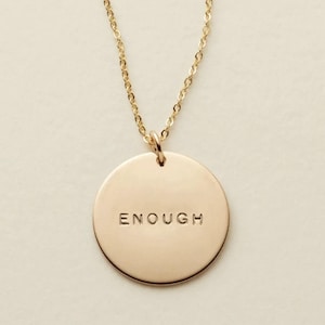 May include: A gold-coloured necklace featuring a circular pendant. The pendant is inscribed with the word "ENOUGH". The chain and pendant share a matching gold tone, resulting in a simple and elegant design.