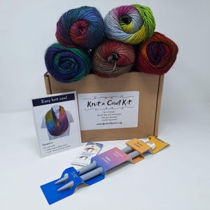 May include: A Knit a Cowl Kit in a cardboard box with five balls of multi-coloured yarn in shades of blue, green, purple, and brown. Includes a printed instruction card, a small packet of sewing needles, and a pair of 25 cm 12 mm knitting needles.