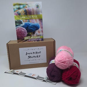 May include: A brown cardboard box with the text "Learn to Knit Starter Kit" on the front. The box contains three balls of yarn in pink, red, and purple, and a pair of knitting needles. The box also includes a booklet titled "Learn to Knit" with the text "Enjoy! Mindful and Relaxing Try our basic patterns".
