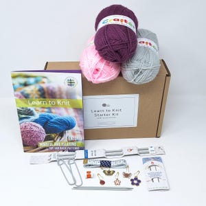 May include: A knitting starter kit in a cardboard box with three yarn balls in purple, pink, and grey. Includes a Learn to Knit instruction booklet, 4 mm knitting needles, a measuring tape, two stitch holders, and various decorative stitch markers.
