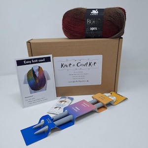 May include: A knitting kit containing a ball of Cygnet Boho Spirit yarn, knitting needles, and a pattern for a cowl. The kit is packaged in a brown cardboard box. The yarn is a blend of red, brown, and tan colours.