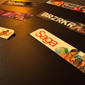 Puede incluir: Una colección de portadas de novelas gráficas de cómics con títulos como "Venom", "Saga", "BRZRKR" y "Strange Academy".