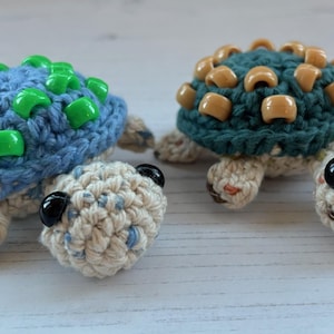 May include: Two handmade crochet turtle figurines. One has a blue shell with green beads, the other has a teal shell with tan beads. Both have off-white bodies, black eyes, and small feet.
